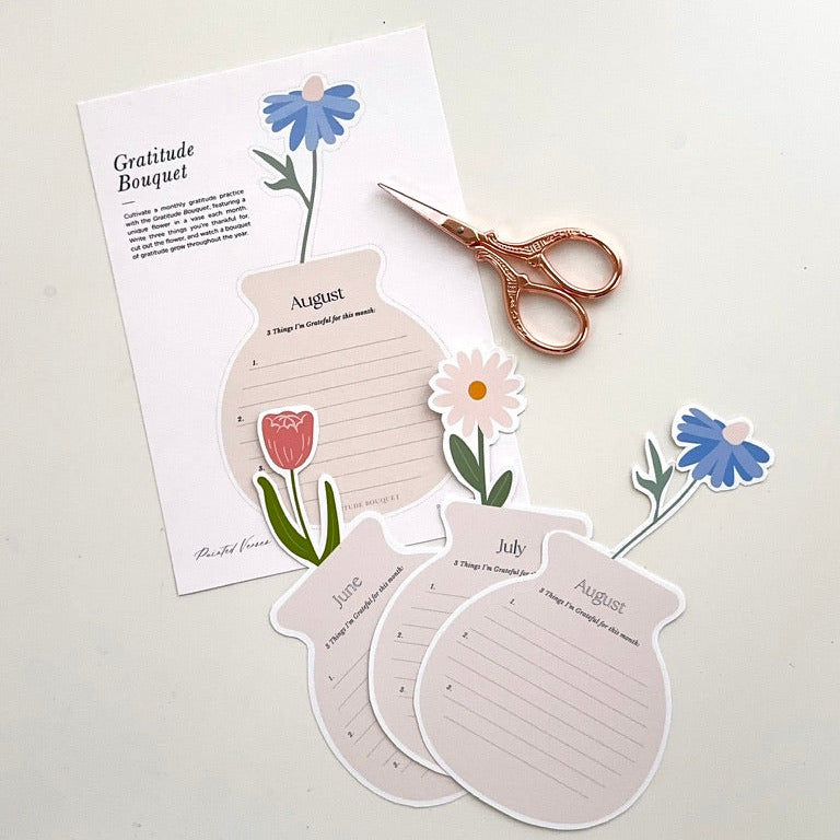 A flat-lay of a gratitude journal page labeled 'Gratitude Bouquet' with illustrated flowers, three monthly cutout pages, and rose gold scissors on a light neutral background.